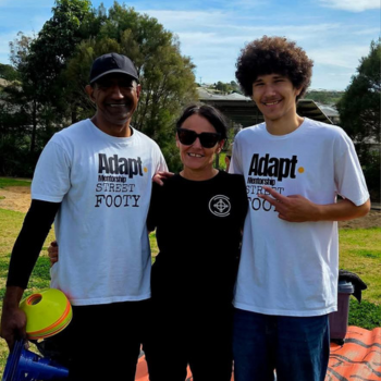 Three people smiling outdoors at a community sports event, two wearing Adapt Mentorship Street Footy t-shirts, with sports equipment and green parkland behind them